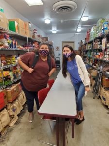 A photo of two masked females in the middle of a food pantry area. The female on the left is Maira Munoz, the person featured in this post.