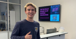 A photo of Grant Berger in the reception area of the RELLIS Building #2. There is a screen in the background with the RELLIS CO-OP advertisement on the screen.
