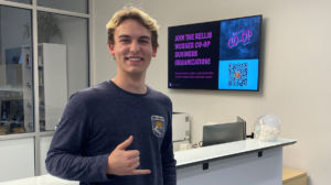 A photo of Grant Berger in the reception area of the RELLIS Building #2. There is a screen in the background with the RELLIS CO-OP advertisement on the screen.